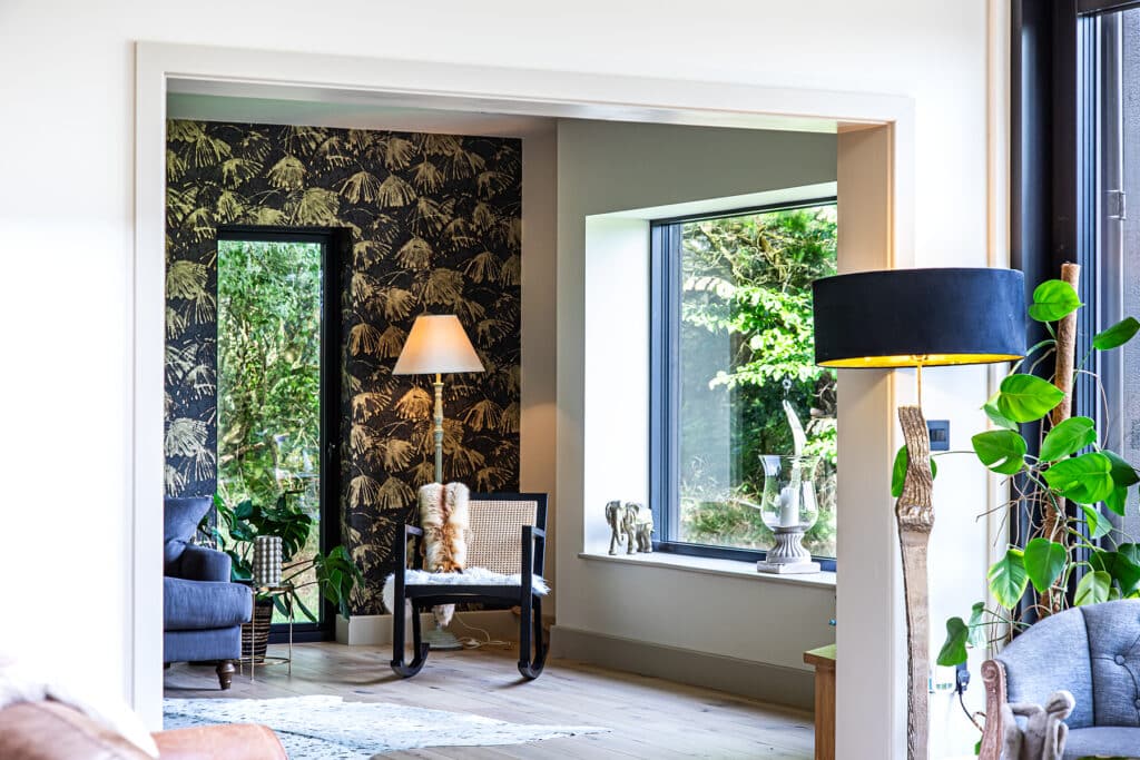 A modern living room with large windows, a patterned feature wall, armchairs, plants, and a standard lamp. Natural light fills the space, highlighting the décor.