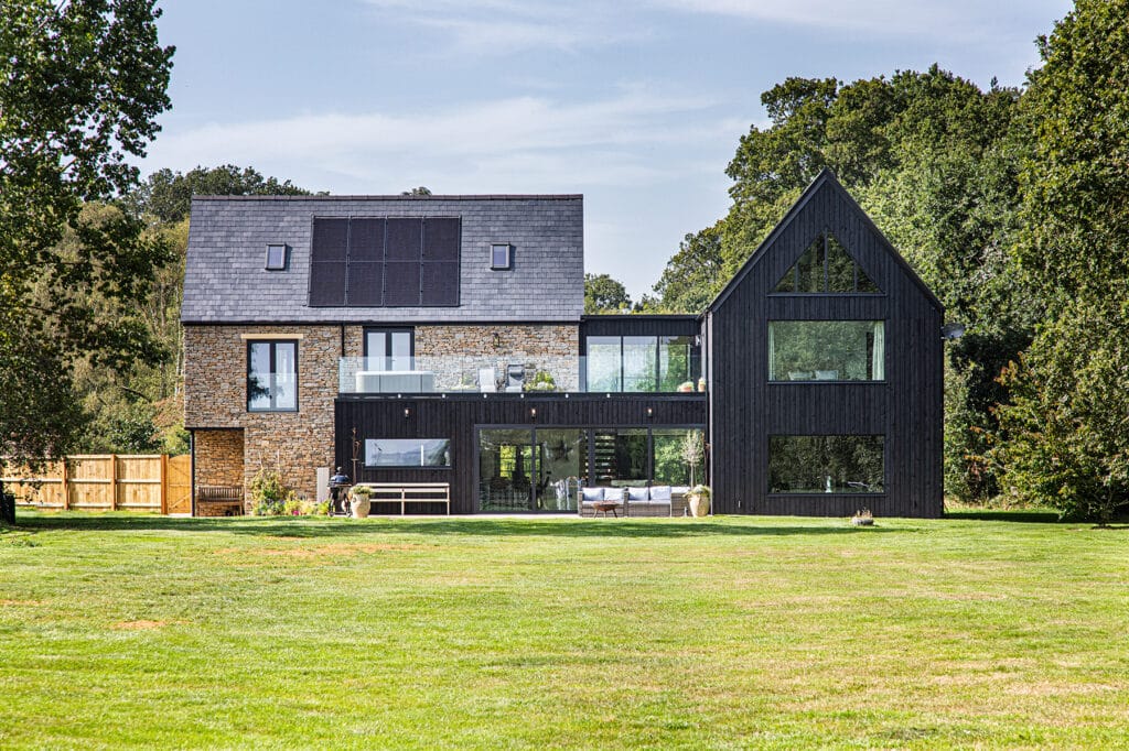 Modern two-storey house with brick and black timber exterior, large windows, solar panels on the roof, and a spacious lawn in the foreground.