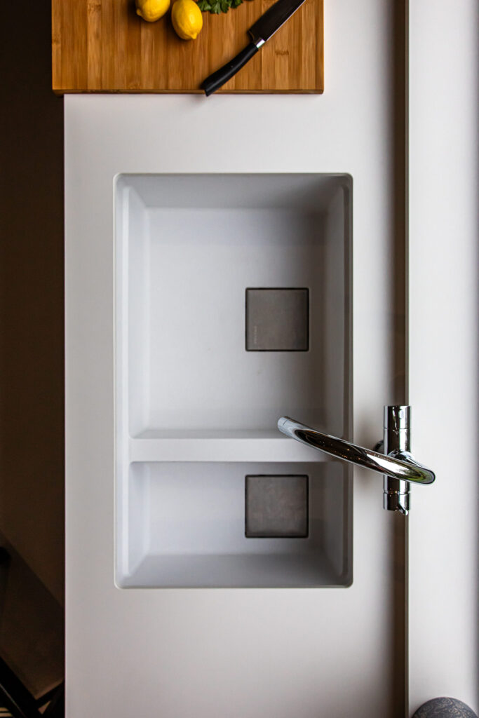 Overhead view of a modern white double kitchen sink with a chrome tap; a chopping board with lemons and a knife is partially visible at the top.