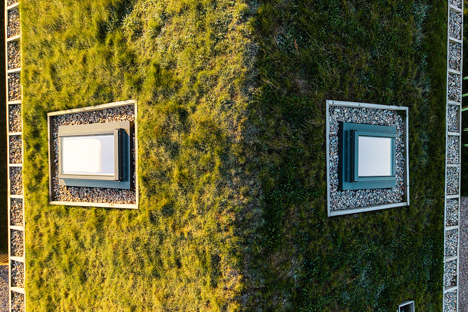 A building corner with a green roof, featuring two square windows surrounded by gravel, covered by dense grass and bordered by concrete tiles.