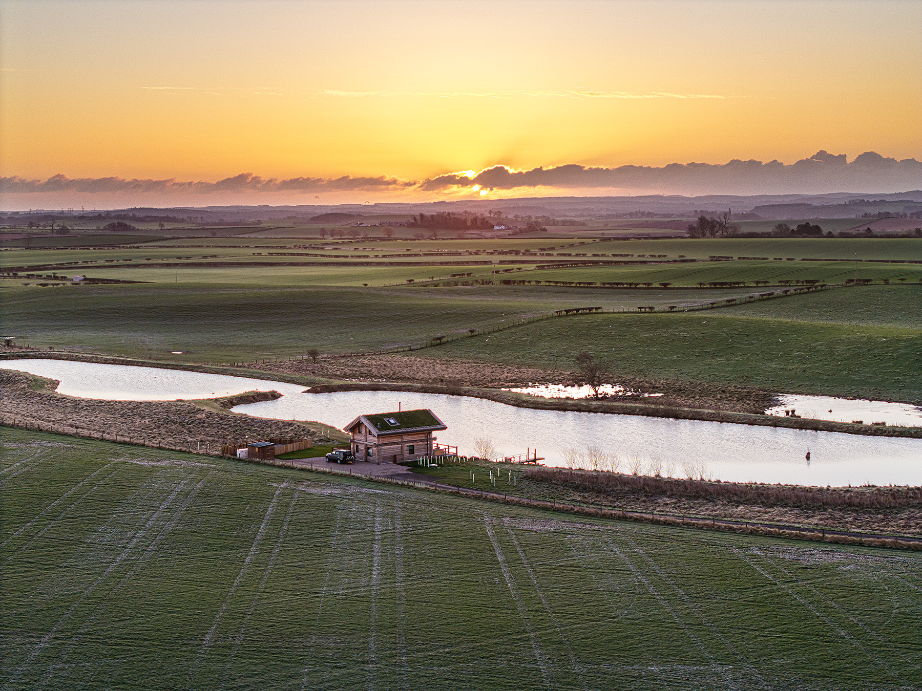 A small house sits beside a narrow river in a wide, open field at sunrise, with the sun just above the horizon and light mist over the distant landscape.