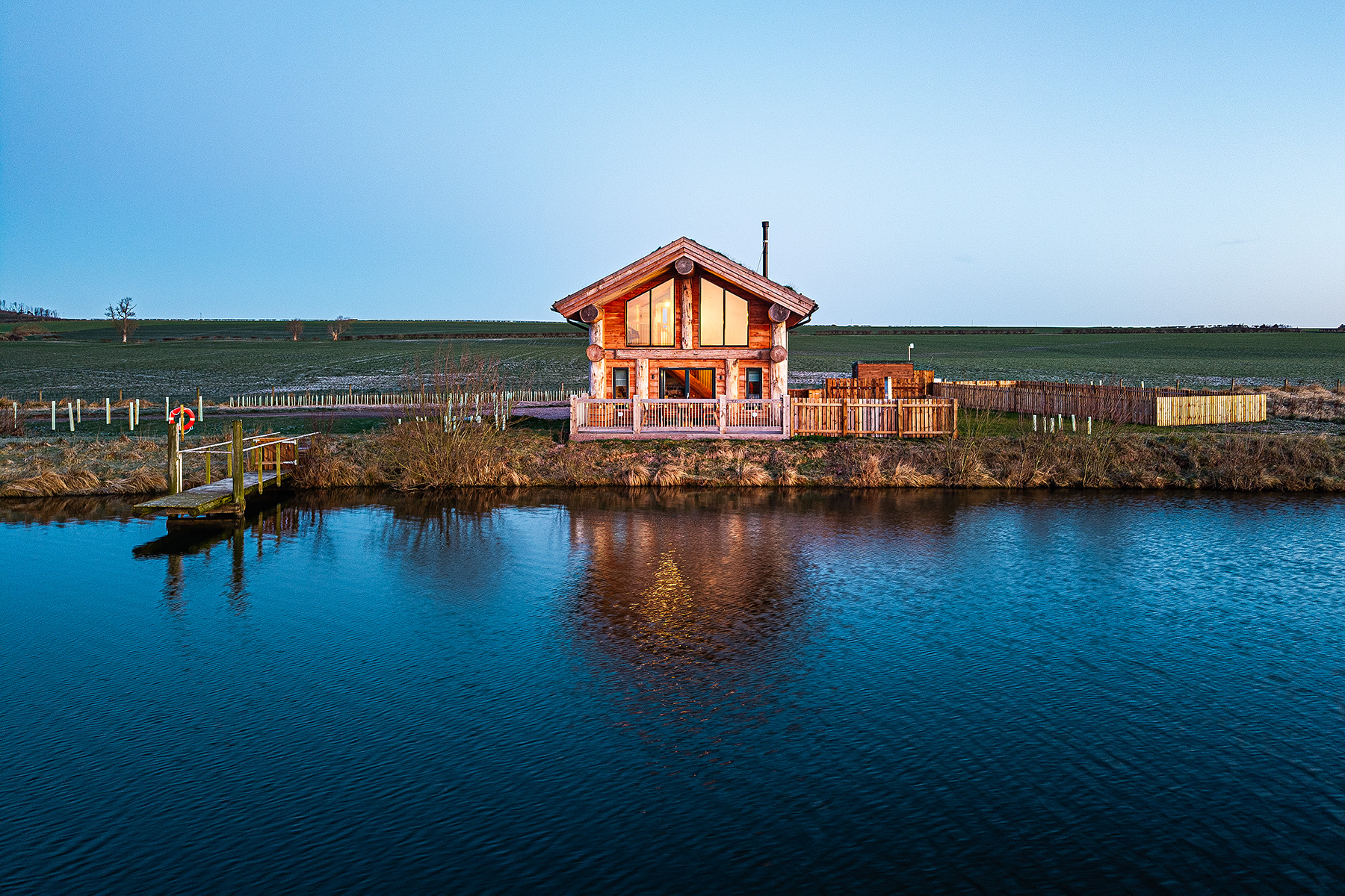 A wooden cabin stands by the edge of a calm lake, reflecting in the water, with open fields and a clear sky in the background.