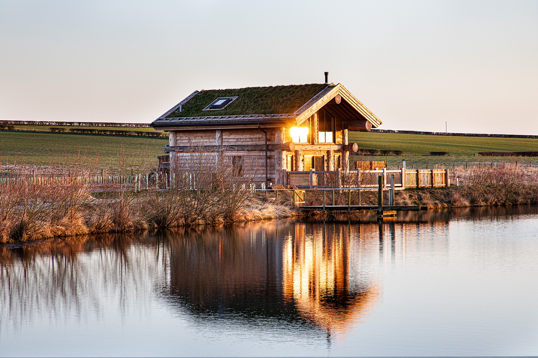 A wooden cabin with a grass roof is reflected in a calm pond, with fields and a clear sky in the background at sunset.
