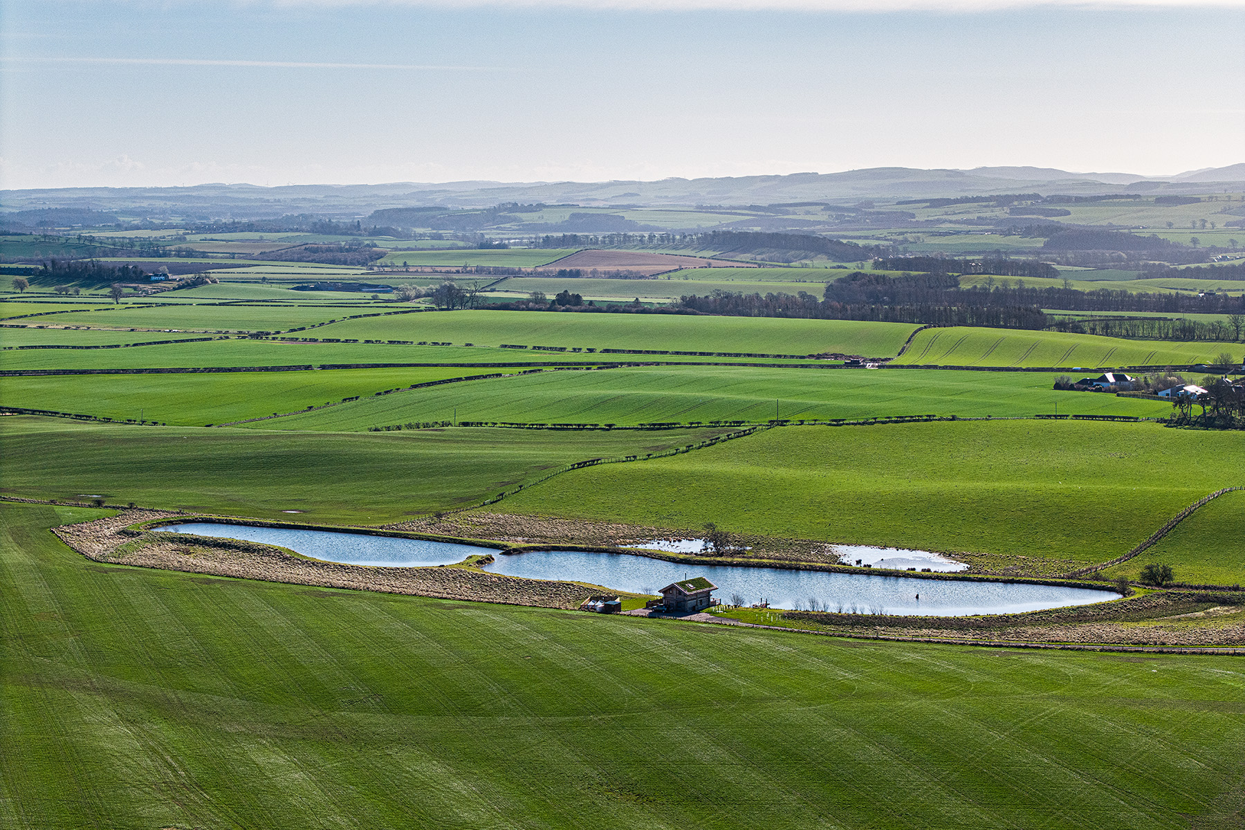 Aerial view of a rural landscape with green fields, a small pond, and a building near the pond, with rolling hills visible in the distance under a clear sky.