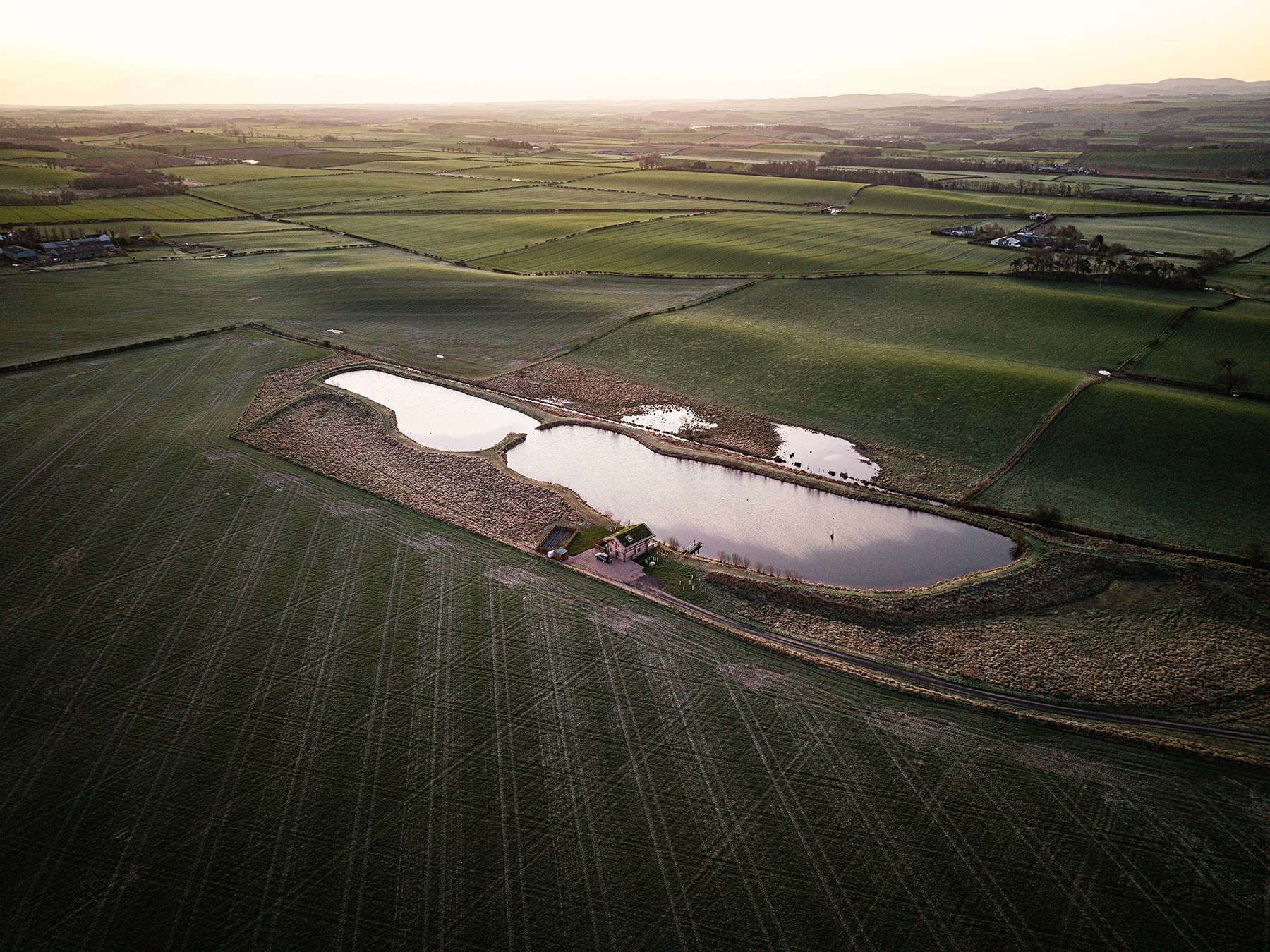Aerial view of a small lake surrounded by grassy fields and farmland at sunset, with a building near the water and long shadows cast across the landscape.
