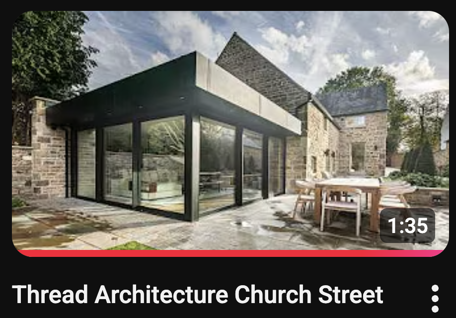 Modern glass extension with black frame attached to a traditional stone house; outdoor patio with dining table and chairs visible in the foreground.