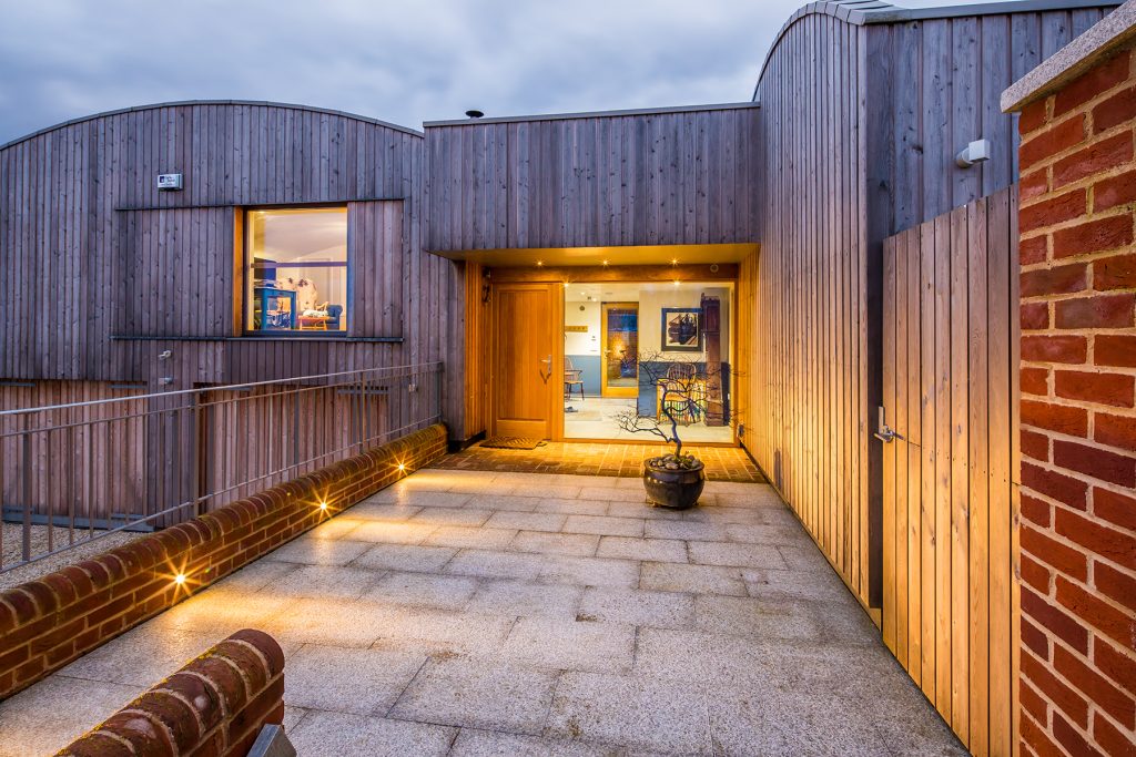 Modern wood-clad house with large windows, lit entrance, outdoor planter, and brick and metal railing along the walkway at twilight. Photograph by Dug Wilders.