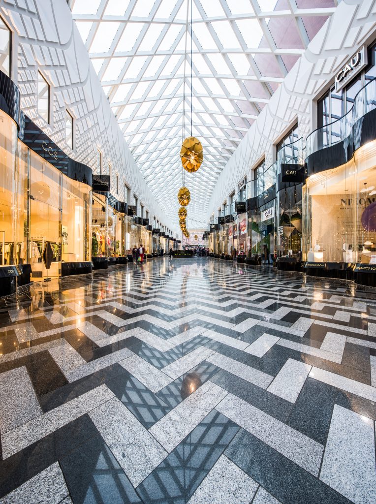 A modern indoor shopping centre with geometric patterned flooring, glass shopfronts, a high glass ceiling, and large gold decorations hanging overhead. Photograph by Dug Wilders.