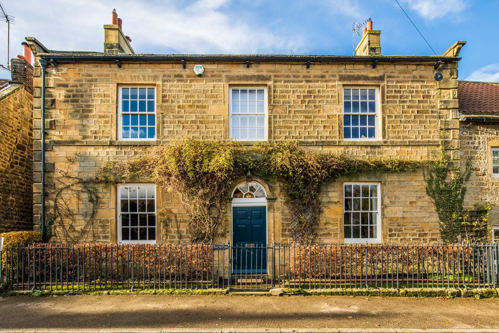 A stone two-storey house with blue door, white-trimmed windows, and vines growing on the façade, behind a black metal fence. Photograph by Dug Wilders.