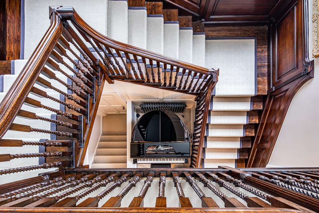 A spiral wooden staircase with white carpeted steps surrounds a black grand piano on the floor below, viewed from above. Photograph by Dug Wilders.