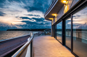 A modern building with large windows and a curved balcony overlooks the sea at sunset, with clouds and a distant coastline visible.