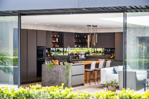 Modern open-plan kitchen with dark cupboards, island with bar stools, built-in appliances, pendant lights, and large windows overlooking a garden.
