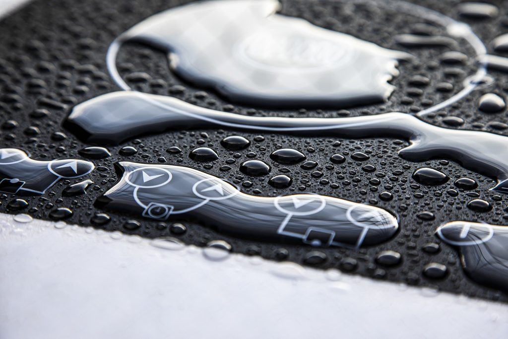 Close-up of water droplets on a black surface with white directional arrow symbols, suggesting moisture on an electronic control panel. Photograph by Dug Wilders.
