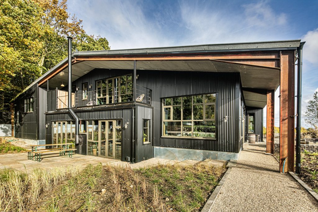 A modern black metal building with large windows, outdoor seating, and a gravel path, set amid greenery and trees under a partly cloudy sky. Photograph by Dug Wilders.