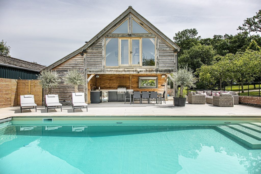 Modern outdoor dining and lounge area with a wooden barn structure, patio furniture, and a swimming pool in the foreground. Photograph by Dug Wilders.