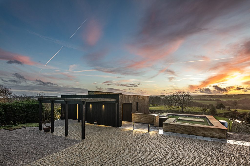 Modern house with wood and dark exterior finishes, a hot tub on a stone patio, and scenic countryside views at sunset with a colourful sky. Photograph by Dug Wilders.