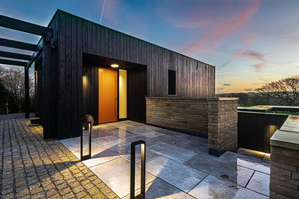 Modern house entrance at sunset with black timber cladding, a bright orange door, brick walls, and illuminated pathway lights. Photograph by Dug Wilders.