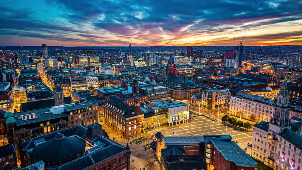 Aerial view of Leeds at sunset, with illuminated buildings, a central square, and a dramatic sky with clouds and fading sunlight