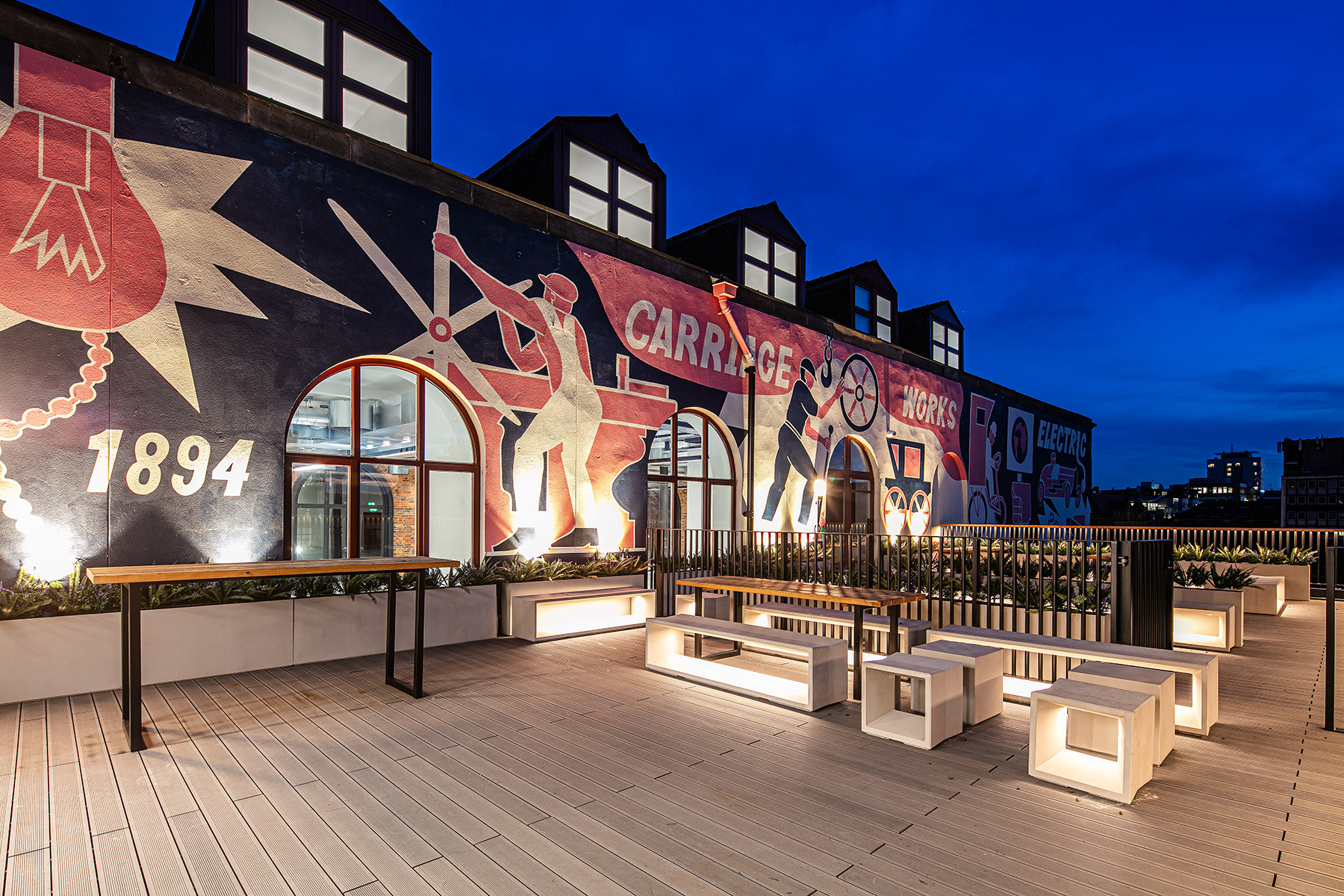 A rooftop terrace at dusk features modern seating and tables, illuminated by lights, with a large mural depicting industrial-themed artwork and the text CARRIAGE WORKS 1894.