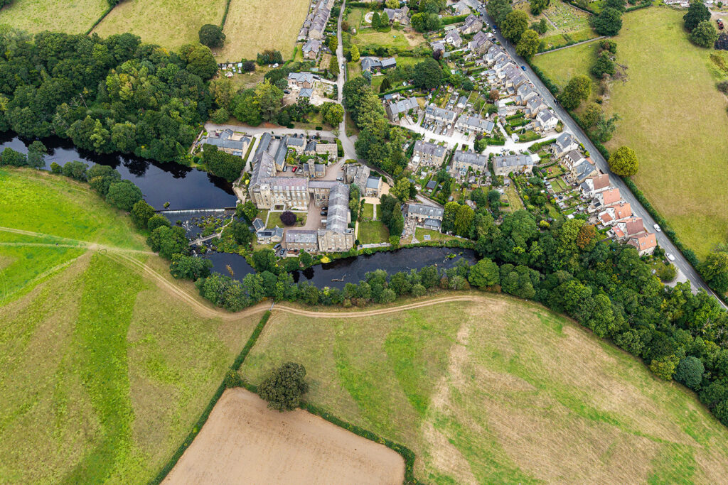 Aerial view of a small village with houses, buildings, trees, and a river surrounded by fields and farmland. Looking over Bamford and the river Derwent