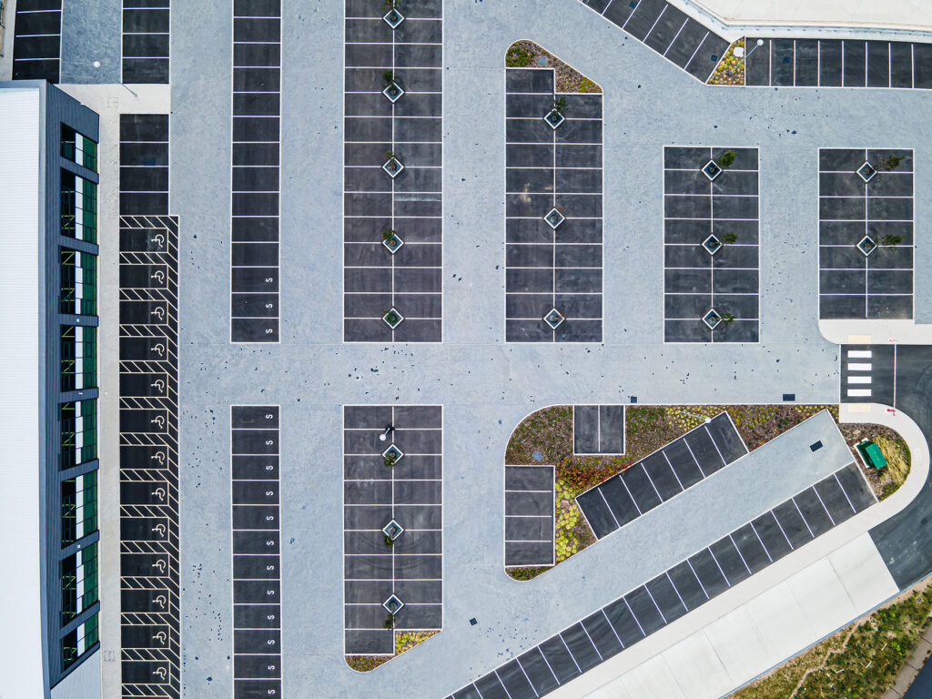 Aerial view of an empty car park with marked spaces, some landscaped islands, and a large building along the left edge.