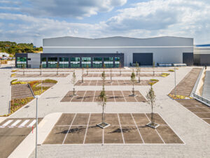 Large, modern warehouse building with multiple garage doors and an empty car park lined with young trees under a partly cloudy sky.