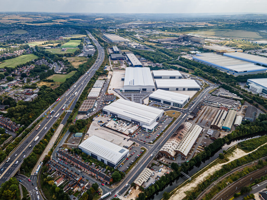Aerial view of an industrial estate with large warehouses, surrounded by roads, motorways, residential areas, and agricultural land.
