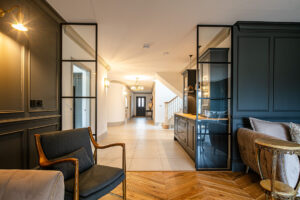 Modern interior featuring a dark feature wall, glass partitions, an armchair, a small round table, and an open hallway leading to a staircase and front door. Photograph by Dug Wilders.