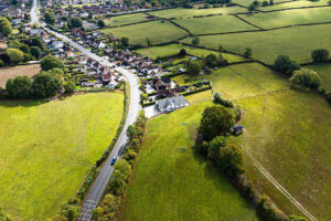 Aerial view of a rural area with houses, a road, open green fields, and scattered trees on a sunny day.
