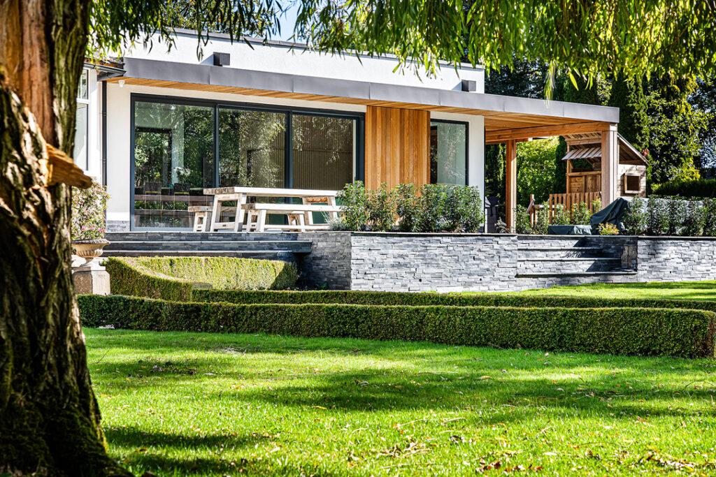 Modern house, large glass doors, wooden accents, patio, picnic table, stone steps, neatly landscaped garden, trimmed hedges, lawn.