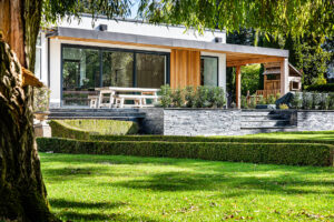 Modern house, large glass doors, wooden accents, patio, picnic table, stone steps, neatly landscaped garden, trimmed hedges, lawn.