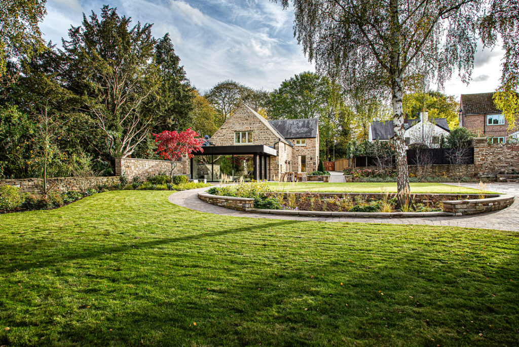A stone house with large windows sits amid a spacious, well-manicured lawn bordered by trees and a curved stone wall.