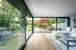 Modern living room with large glass sliding doors open to a patio and garden area; features a corner sofa, coffee table, and views of trees and stone walls outside.
