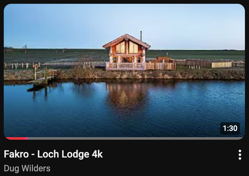 A wooden lodge sits by the edge of a calm loch in Scotland, with open fields in the background under a clear sky.