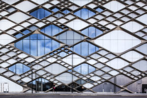 A person walks in front of a modern building facade with geometric, diamond-shaped metal patterns and large glass windows reflecting the sky.