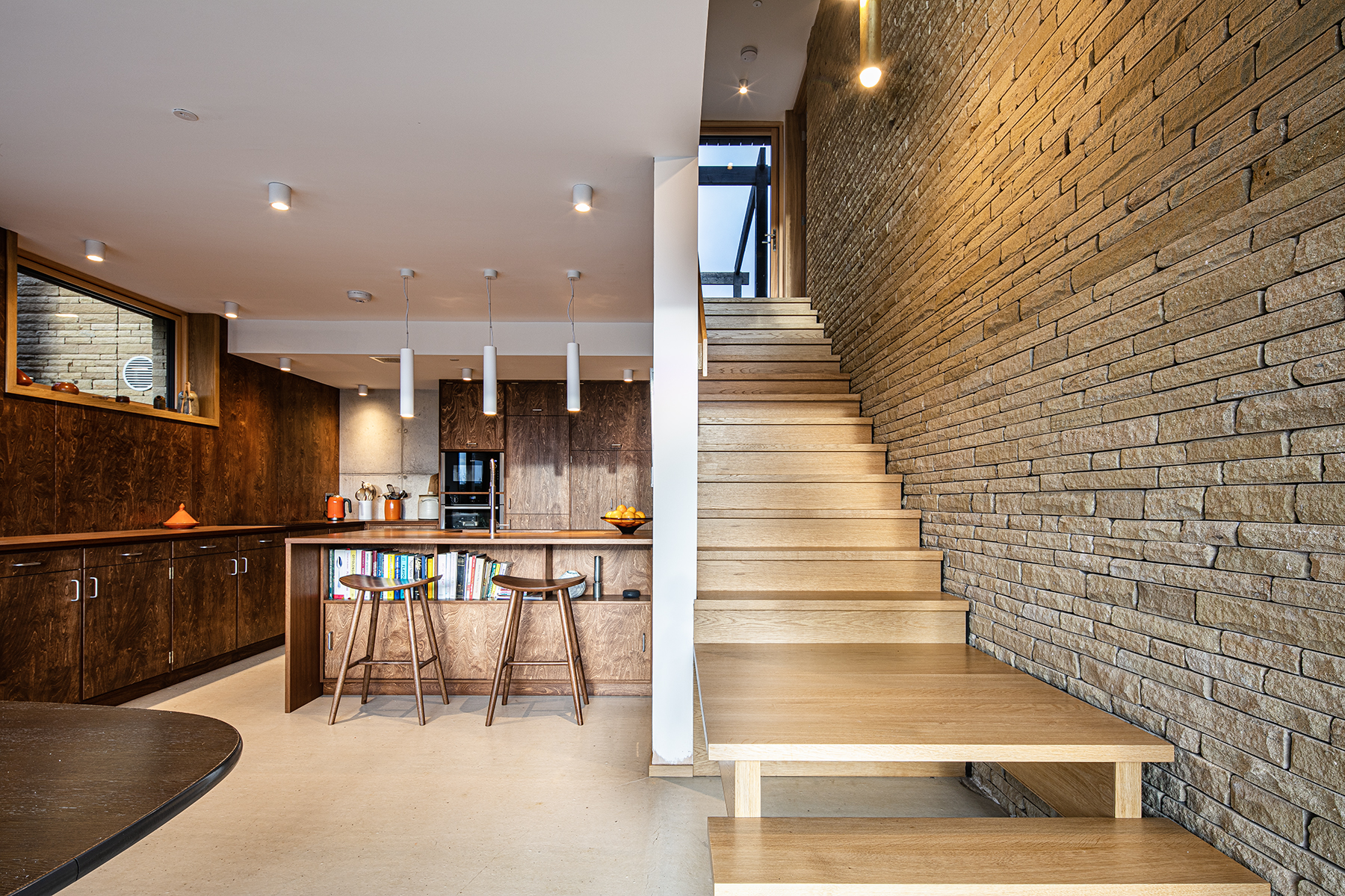 Modern kitchen with wooden cupboards, an island with stools, open shelving, and adjoining wooden staircase against a textured brick wall—captured by an architectural photographer inspired by the natural textures and refined style of the Peak District.